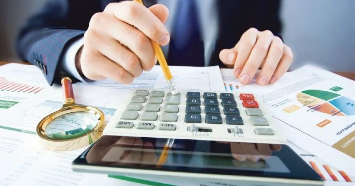 Cineva face calcule la un calculator, lângă o lupă și niște grafice economice
